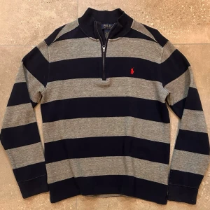 Randig stickad tröja från Polo Ralph Lauren - Mörkblå och grå stickad tröja med breda ränder från Polo Ralph Lauren. Tröjan har halv dragkedja vid halsen, lång ärm och en röd broderad logga på bröstet. Tillverkad i mjukt material som passar perfekt till en avslappnad stil.