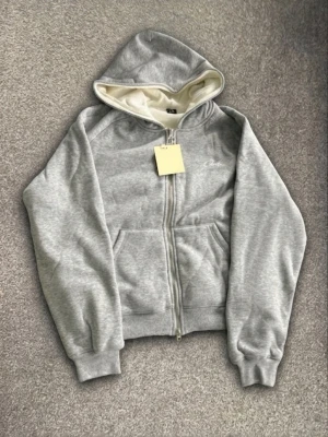 Ljusgrå hoodie med dragkedja - Säljer en ljusgrå hoodie med dragkedja och huva. Hoodien har fickor framtill och en liten vit broderad text på bröstet där det står 'Cyntre'. Insidan av huvan är vit och materialet känns mjukt och bekvämt, perfekt för chill dagar.