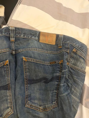 Blå jeans från Nudie Jeans - Säljer ett par klassiska blå jeans från Nudie Jeans med snygga slitningar och kontrastsömmar. Jeansen har raka ben, fem fickor och tydlig vågdetalj på bakfickorna. Tillverkade i denim med cool tvätt och detaljer i orange. På sista bilden kan man se att jeansen har ett hål mellan benen. Därav priset.