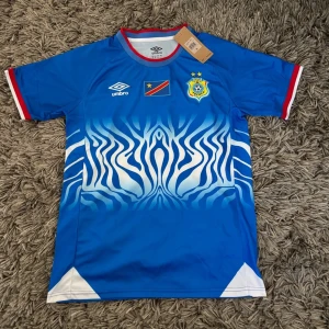 Blå ECOF fotbollströja från Umbro - Snygg blå fotbollströja från Umbro med ECOF-märke och Kongos flagga på bröstet. Tröjan har ett unikt zebramönster i vitt och blått framtill, röda och vita detaljer vid ärmslut och hals. Perfekt för dig som gillar sportig stil.