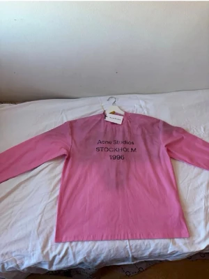 Rosa långärmad t-shirt Acne Studios - Säljer en rosa långärmad t-shirt från Acne Studios med trycket 'Acne Studios STOCKHOLM 1996' på bröstet. T-shirten har en klassisk passform och är tillverkad i mjuk bomull. Perfekt för dig som gillar stilrena och trendiga plagg.