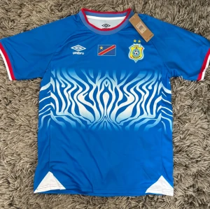 Blå Umbro DR Kongo fotbollströja - Säljer en blå fotbollströja från Umbro med DR Kongos flagga och förbundsmärke på bröstet. Tröjan har ett vitt zebramönster framtill, röda och vita detaljer vid ärmslut och krage samt en sportig passform. Perfekt för match eller träning.