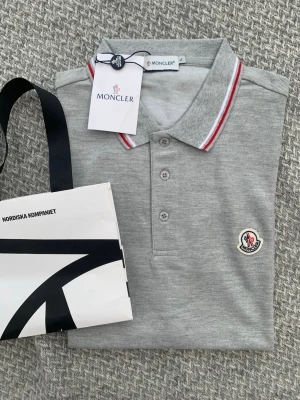 Grå pikétröja från Moncler - Snygg grå pikétröja från Moncler med klassisk krage och knappar framtill. Kragen har vita och röda ränder som ger en sportig touch. Moncler-logga är broderad på bröstet. Perfekt till jeans eller shorts.