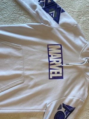 Vit Marvel hoodie med lila tryck - Snygg vit hoodie med stort lila Marvel-tryck på bröstet och coola grafiska detaljer på ärmarna. Baksidan har ett stort motiv med två Marvel-karaktärer i svartvitt och lila. Perfekt för dig som älskar superhjältar och streetstyle.