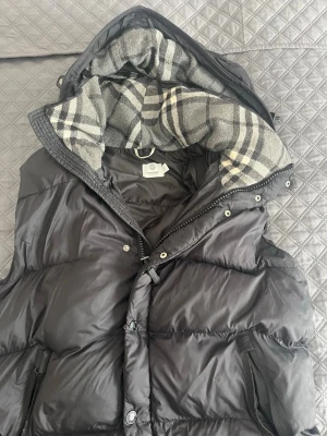 Burberry väst Svart dunväst med huva  - Snygg svart dunväst med dragkedja och knappar framtill. Västen har en stor huva fodrad med grått och vitt rutigt tyg som ger en cool kontrast. Två fickor med dragkedja på sidorna och en sportig look. Perfekt för lager-på-lager-stil. Västens vänstra fika är trasig men utöver det så finns det inga problem med västen. Har bärt den 3 gånger och den är i ett väldigt bra skick. Storleken är M, men västen är stor i storleken så det känns mest som en L/XL. Priset kan såklart diskuteras 
