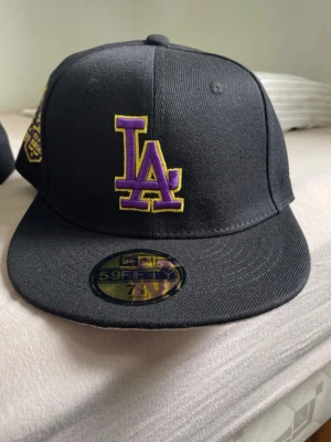 Svart LA keps med lila detaljer - Säljer en svart New Era 59FIFTY keps med LA-logga i lila och gul brodyr framtill. Klassisk flat brim och broderad New Era-logga på sidan. Perfekt för dig som vill ha en stilren och sportig look.