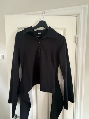 Svart skjorta med knytband - Säljer en svart skjorta med långa ärmar och unika knytband baktill. Skjortan har klassisk krage och knäppning med knappar. Perfekt för dig som vill ha en stilren men ändå edgy look.