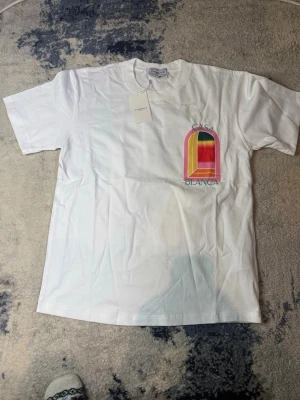 Vit Casa Blanca t-shirt med färgglatt tryck - Vit t-shirt från Casa Blanca med ett stort färgglatt grafiskt tryck i rosa, gult, grönt och rött både fram och bak. T-shirten har klassisk passform och rund hals. Perfekt för dig som gillar statement-plagg och vill sticka ut. Väskan kommer också med