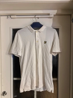 Vit Piké från Lyle & scott - Klassisk vit pikétröja med krage och tre knappar framtill. Tröjan har korta ärmar och en liten broderad logga i gult och svart på bröstet. Tillverkad i mjuk bomull, perfekt för en clean och enkel stil.