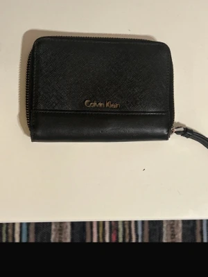 Plånbok Calvin Klein - Snygg svart plånbok från Calvin Klein i läder med struktur. Plånboken har dragkedja runtom, flera kortfack och ett myntfack med dragkedja. Calvin Klein-logga i metall på framsidan och insidan. Perfekt storlek för fickan eller väskan.