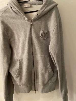 Moncler kofta  - Säljer en grå hoodie med dragkedja och broderad logga på bröstet. Hoodien har huva, fickor framtill och ribbade muddar vid ärmar och nederkant. Perfekt för en chill och avslappnad stil. Materialet känns mjukt och bekvämt.