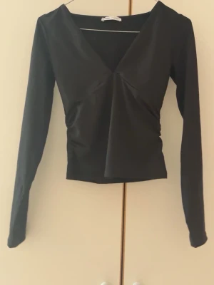 Svart långärmad v-ringad topp - Snygg svart långärmad topp med v-ringning och lätt drapering framtill. Modellen är figurnära och har en enkel, stilren design som passar till många olika looks. Perfekt för dig som gillar minimalistisk stil.