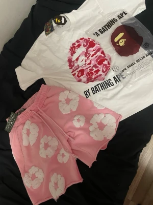Denim tears/bape - Säljer ett par rosa shorts med stora vita blommor. De har elastisk midja och avslappnad passform, perfekta för sommardagar. Materialet känns mjukt och bekvämt, och shortsen har en cool streetwear-vibe.