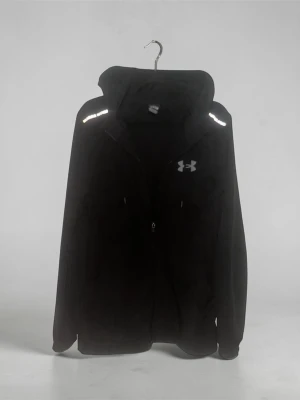Under armour jacka - Svart vindjacka från Under Armour med dragkedja framtill och reflekterande detaljer på axlarna. Jackan har en klassisk huva och logga på bröstet. Tillverkad i lätt och smidigt material som skyddar mot vind. Perfekt för en sportig och stilren look. Storlek S. Ett pyttelitet hål över höger ficka som inte syns när man har på sig den. 