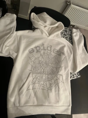 Spider hoodie - Säljer en vit hoodie med huva och stor ficka framtill. På bröstet står det 'spider' i glittrande strass och ett stort spindelnät i strass täcker framsidan. Hoodien har ribbade muddar och är i mjukt material, perfekt för dig som gillar statement-plagg.