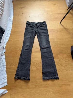 Gråa bootcut jeans med låg midja - Säljer ett par grå bootcut jeans med låg midja och dubbla knappar i linningen. Jeansen har klassiska fem fickor och råa kanter nertill. Materialet är denim med lätt stretch för skön passform. Perfekta för en avslappnad och trendig look. Uppsprättade där nere