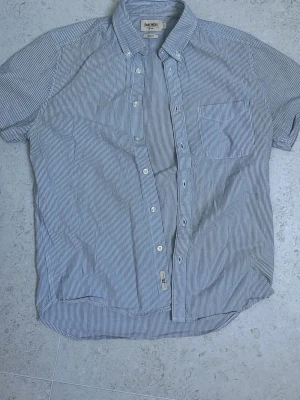 Blårandig kortärmad skjorta East West  - Säljer en blåvitrandig kortärmad skjorta från East West i slim fit. Skjortan har klassisk krage med knappar, bröstficka och stängs med vita knappar framtill. Perfekt till sommaren och tillverkad i ett lätt bomullstyg. Storlek M
