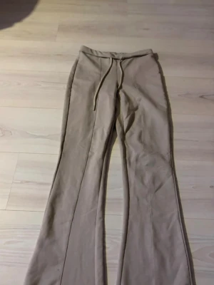Beiga utsvängda mjukisbyxor - Säljer ett par beiga mjukisbyxor med utsvängda ben och dragsko i midjan. Byxorna har en enkel design utan synliga loggor och är gjorda i ett mjukt, stretchigt material som känns skönt mot huden. Perfekta för en avslappnad och chill stil.