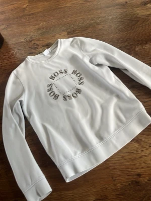 Vit sweatshirt från Hugo Boss - Snygg vit sweatshirt från Hugo Boss med rund hals och tryck med texten 'BOSS' i ett fyrkantigt mönster på bröstet. Tröjan har långa ärmar och är gjord i mjukt bomullsmaterial som känns skönt mot huden. Perfekt för en clean och stilren look.