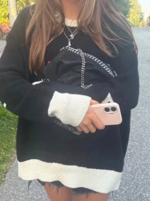 Svart handväska från Stella McCartney - Snygg svart handväska från Stella McCartney med kedjedetaljer i silver och grov söm längs kanterna. Nypris: 7000-10000kr. Väskan har en mjuk, mockaliknande yta och en rund metallbricka med logotyp. Perfekt accessoar för att lyfta din outfit.