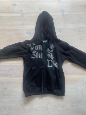 Svart hoodie från Vonstudio XS - Säljer en svart hoodie från Vonstudio i storlek XS. Hoodien har dragkedja, stor huva och fickor framtill. Tryck i vitt med text och en ängel på bröstet ger en edgy vibe. Perfekt för dig som gillar streetwear och vill sticka ut.