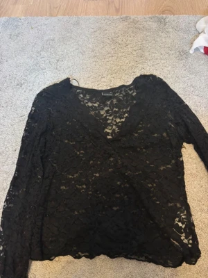 Svart spetsblus med v-ringning - Säljer en svart blus i spets med v-ringad hals och långa ärmar. Blusen har ett transparent, blommigt spetsmönster som ger en snygg och trendig look. Perfekt att styla med ett linne under eller som den är för en mer vågad outfit.