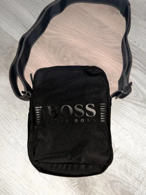 Svart axelväska från Hugo Boss - Snygg svart axelväska från Hugo Boss med justerbar axelrem och dragkedja. Väskan har en stilren design med BOSS-logga framtill och på dragkedjan. Perfekt storlek för det viktigaste och enkel att matcha till olika outfits.