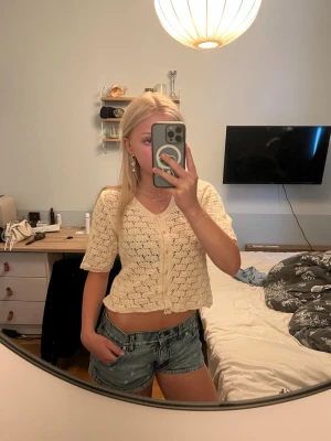Beige croppad stickad topp med knappar - Söt croppad topp i beige stickat material med korta ärmar och v-ringning. Toppen har dekorativa knappar framtill och ett luftigt, hålmönstrat tyg som ger en somrig vibe. Perfekt att styla med jeansshorts eller kjol.