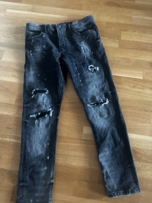 Svarta slitna jeans Valére Milano - Säljer ett par svarta tapered fit jeans från Valére Milano med slitningar och hål på båda benen. Jeansen har fem fickor och coolt tvättad look med distressed detaljer. Perfekta för dig som gillar streetstyle och en avslappnad vibe.