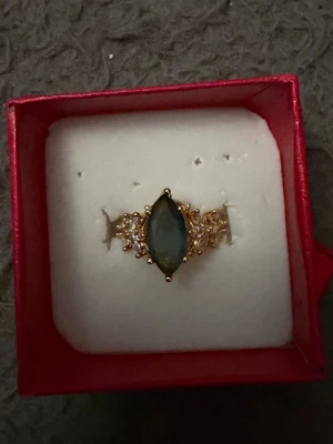 Guldfärgad ring med blå sten - Snygg guldfärgad ring med en avlång, mörkblå sten i mitten och små gnistrande detaljer på sidorna. Ringen har en elegant och unik design som verkligen sticker ut. Perfekt för dig som vill ha något speciellt till din stil.