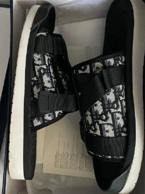 Svarta sandaler  - Säljer ett par svarta sandaler med klassiskt monogrammönster i vitt och svart. Sandalerna har breda textilband med justerbara spännen och en vit, tjock sula för extra komfort. Perfekta för dig som vill ha en trendig och lyxig look.