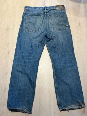 Blå raka jeans från G-Star RAW - Säljer ett par klassiska blå jeans från G-Star RAW med raka ben och två bakfickor. Jeansen har en snygg tvättad look och detaljer som läderpatch och G-Star RAW-märke ovanför fickan. Perfekta för en avslappnad stil. Storlek 34/34