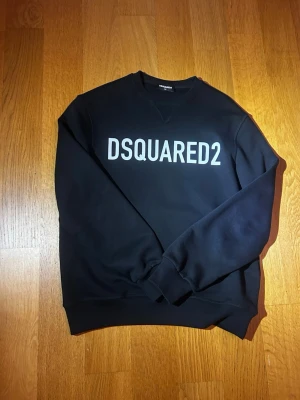 Svart hoodie från DSQUARED2 - Snygg svart hoodie från DSQUARED2 med stor vit logga framtill. Klassisk modell med huva och ribbade muddar vid ärmslut och nederkant. Perfekt för dig som gillar streetwear och vill ha en stilren look. Materialet känns mjukt och bekvämt.