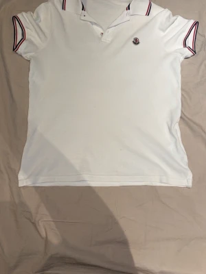 Vit Moncler piké med rödblå detaljer - Snygg vit pikétröja från Moncler med klassisk krage och rödblå ränder på krage och ärmslut. Tröjan har Moncler-logga på bröstet och är tillverkad i mjuk bomull. Perfekt för en clean och stilren look.