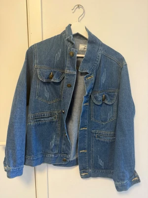 Blå jeansjacka från J.H.L. Jeans - Snygg blå jeansjacka från J.H.L. Jeans med klassisk passform, fyra fickor framtill och slitna detaljer. Jackan har ståkrage, bruna knappar och cool vintagekänsla. Perfekt för dig som vill ha en tidlös denimlook.