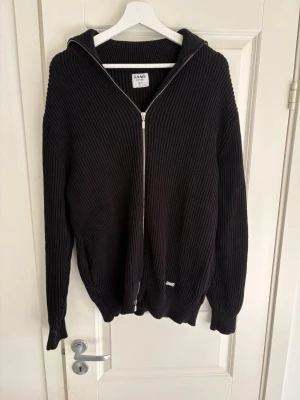 Svart ziptröja med huva från SAMS - Riktigt snygg svart ziptröja från SAMS i grovstickat material. Tröjan har huva, dragkedja hela vägen och ribbade muddar. Perfekt för dig som gillar en enkel men stilren look. Passar till det mesta och är superbekväm.