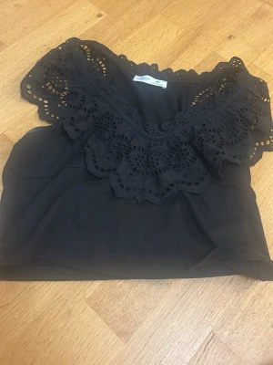 Svart topp med broderad spets - Snygg svart topp med bred broderad spets runt urringningen. Toppen har korta ärmar och är gjord i ett mjukt, tunt tyg som känns skönt mot huden. Perfekt för dig som gillar detaljer och vill ha något extra till din outfit.
