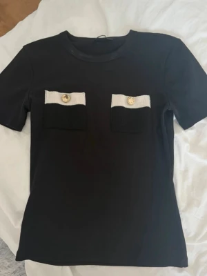 Svart t-shirt med fickdetaljer - Snygg svart t-shirt med två dekorativa fickor framtill. Fickorna har svart och vit ribbad kant samt guldfärgade knappar för en cool kontrast. T-shirten har rund halsringning och korta ärmar. Perfekt för dig som vill ha en unik twist på din outfit.