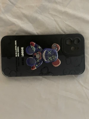 Svart mobilskal med björnmotiv - Snyggt svart mobilskal med ett färgglatt björnmotiv och textdetaljer. Skalet har ett popsocket-liknande grepp i form av en björn och är gjort i tåligt plastmaterial. Passar mobil med dubbla kameror och blixt. Perfekt för dig som vill ha ett unikt och trendigt skal.