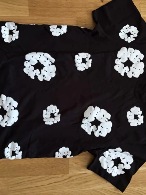 Svart t-shirt med vita blommor - Cool svart t-shirt från Danny Tears i storlek S, dekorerad med stora vita blommönster över hela plagget. T-shirten har rund hals och korta ärmar, perfekt för en avslappnad och trendig stil. Materialet känns mjukt och bekvämt mot huden.