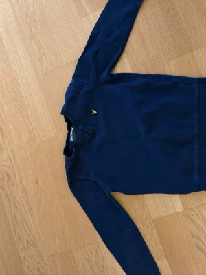Mörkblå sweatshirt från Lyle & Scott - Mörkblå sweatshirt från Lyle & Scott med klassisk gul logga på bröstet. Tröjan har rund halsringning och långa ärmar. Tillverkad i mjukt bomullsmaterial som passar perfekt för kyligare dagar.