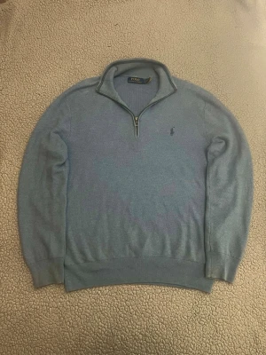 Ljusblå halvzip från Polo Ralph Lauren - Ljusblå stickad tröja från Polo Ralph Lauren med halvzip och broderad logga på bröstet. Tröjan har ribbade muddar vid ärmslut och nederkant samt en hög krage. Perfekt för lager på lager och riktigt snygg till jeans.