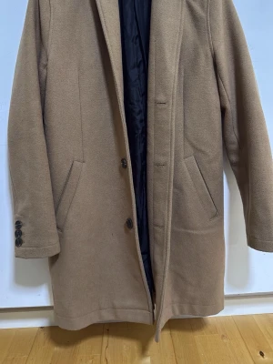 Klassisk beige ullkappa - Stilren beige kappa i ullmix med rak passform och knäppning framtill. Fodrad med svart innerfoder och har två snedställda fickor. Perfekt för kyliga dagar och ger en tidlös look till din outfit.