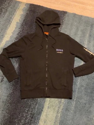 Svart hoodie med tryck från BOSS - Svart hoodie från BOSS med dragkedja framtill, huva med snörning och fickor. Liten BOSS-logga och ljudvågsdetalj på bröstet samt stort grafiskt tryck med texten 'Tune and Play' på ryggen. Perfekt för en avslappnad streetwear-look. Storlek m men är liten i passformen