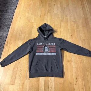 Saint Laurent hoodie - Navy blå  hoodie från Saint Laurent med tryck framtill i vitt och rött, samt texten 'SAINT LAURENT'. Tröjan har huva med snörning och en stor magficka. Tröjan har långa ärmar perfekt för en chill och stilren look.