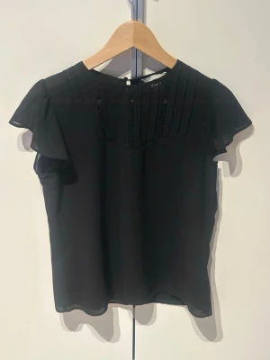 Svart blus med volangärmar - Stilren svart blus i tunt, lätt material med korta volangärmar. Framsidan har dekorativa veck och spetsdetaljer upptill. Blusen har rund halsringning och knapp i nacken. Perfekt att matcha med jeans eller kjol.