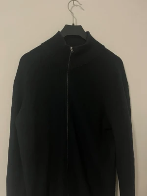 Mörkblå Cardigan från Dressmann - Säljer en mörkblå stickad ziptröja från Dressmann med hög krage och hel dragkedja framtill. Tröjan har en enkel och stilren design, perfekt att slänga på sig över en t-shirt. Materialet är mjukt och bekvämt.
