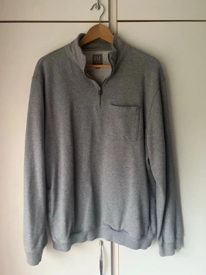 Grå halvzip hoodie från GAP - Säljer en stilren grå halvzip hoodie från GAP. Tröjan har en hög krage med dragkedja, bröstficka och ribbade muddar vid ärmslut och nederkant. Perfekt för en chill och avslappnad look. Materialet är mjukt och skönt bomullsmix.
