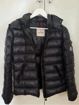 Svart dunjacka från Moncler med huva - Snygg svart dunjacka från Moncler med avtagbar huva och två dragkedjeförsedda fickor framtill. Jackan har quiltad design, diskret Moncler-logga på ärmen och dragkedja framtill. Perfekt för kalla vinterdagar och riktigt stilren look. Den är svart men kan även se lite mörkblå ut ibland.