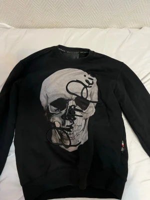 Svart sweatshirt med dödskalletryck - Cool svart sweatshirt med stort dödskalletryck på framsidan och broderad text. Tröjan har rund hals och ribbade muddar vid ärmslut och nederkant. Perfekt för dig som gillar edgy streetstyle och vill sticka ut.